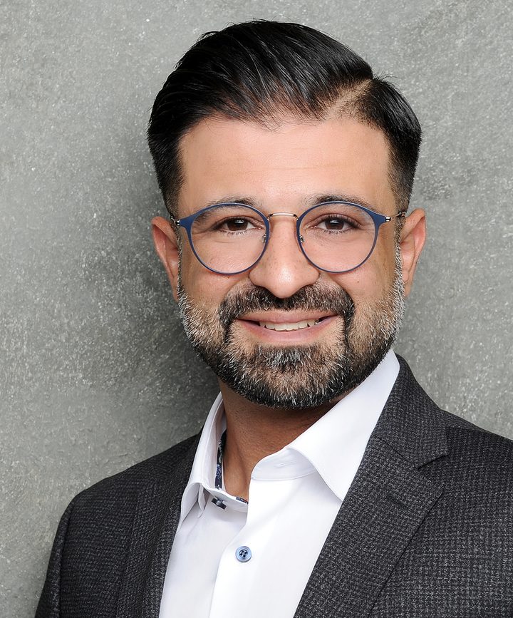 Babak Rabbany mit Brille und dunklem Sakko lächelt in die Kamera, vor grauer Betonwand.
